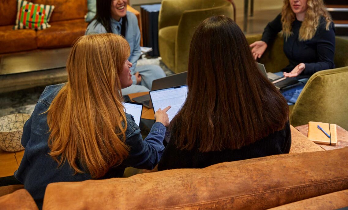 Two Levi Strauss & Co. employees sit on a couch with their backs to the camera. They point at a computer screen on the right-hand employees lap. Two other employees can be seen chatting in the background.