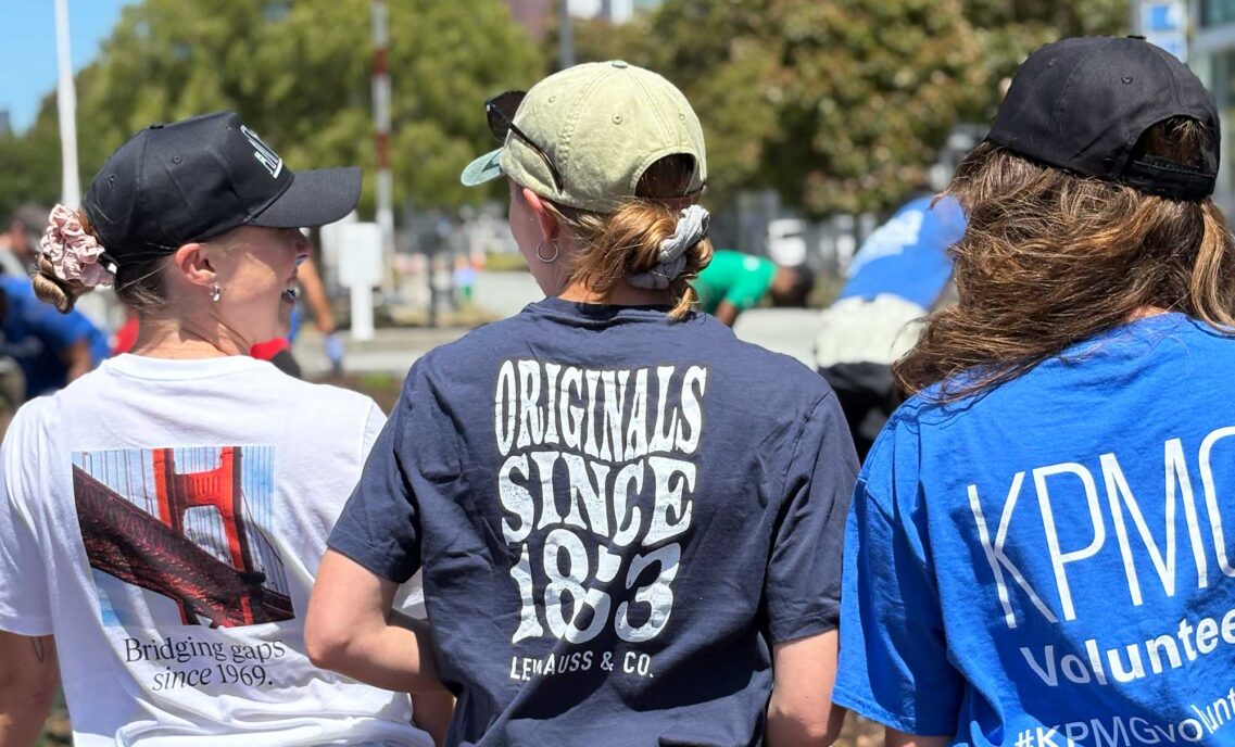 A Gap employee, an LS&Co. employee and KPMG employee stand with their backs to the camera wearing their company volunteer shirts at a Downtown Volunteer Coalition volunteer project