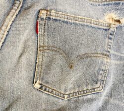 The back pocket of a pair of Levi's® 501® jeans, featuring a red Tab on the top left corner of the pocket, an Arcuate stitch, and a torn hole on the top right corner