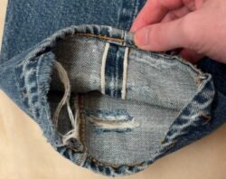 A hand lifts up the bottom cuff of a pair of Levi's® 501® jeans, revealing a white selvedge line against the dark blue indigo color of the denim