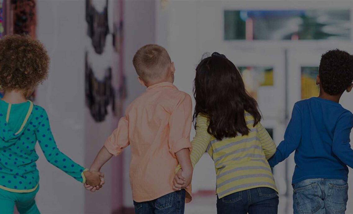 Four children hold hands in a school hallway with their backs to the camera