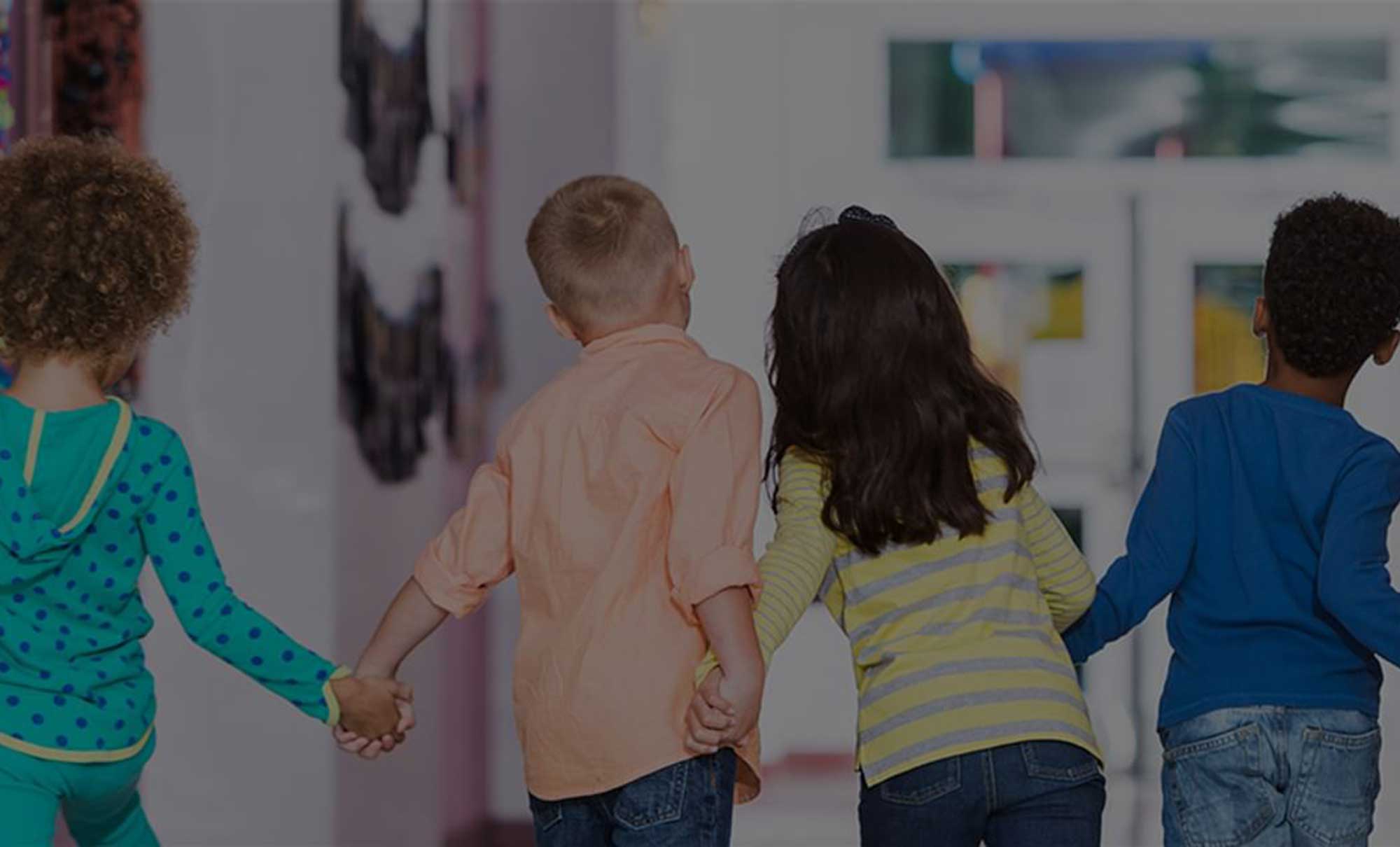 Four children hold hands in a school hallway with their backs to the camera