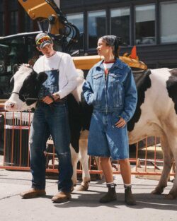 A black and white cow stands behind a person wearing denim overalls over a white long sleeve top next to a person wearing a denim jacket and long denim shorts. The group stands in front of a construction site.