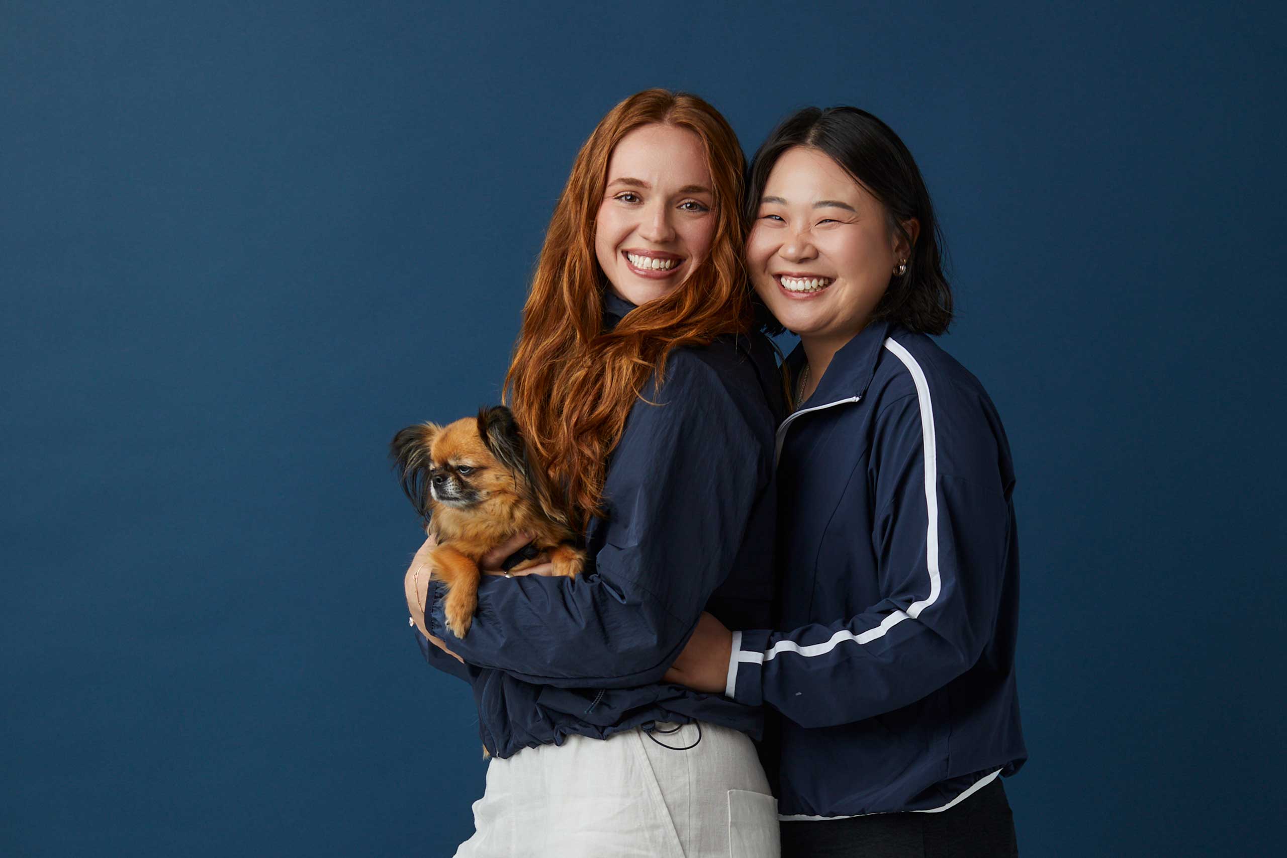 Two LS&Co. employees from Beyond Yoga pose next to each other in front of a blue background. The employee on the left smiles while holding a small dog, while the employee on the right leans in and smiles