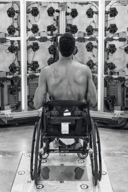 A black and white image of the back of Antwan Tolliver, founder of FREEDOM IS FLY, sitting in his wheelchair being scanned by cameras to create a mannequin cast made from a 3D scan.