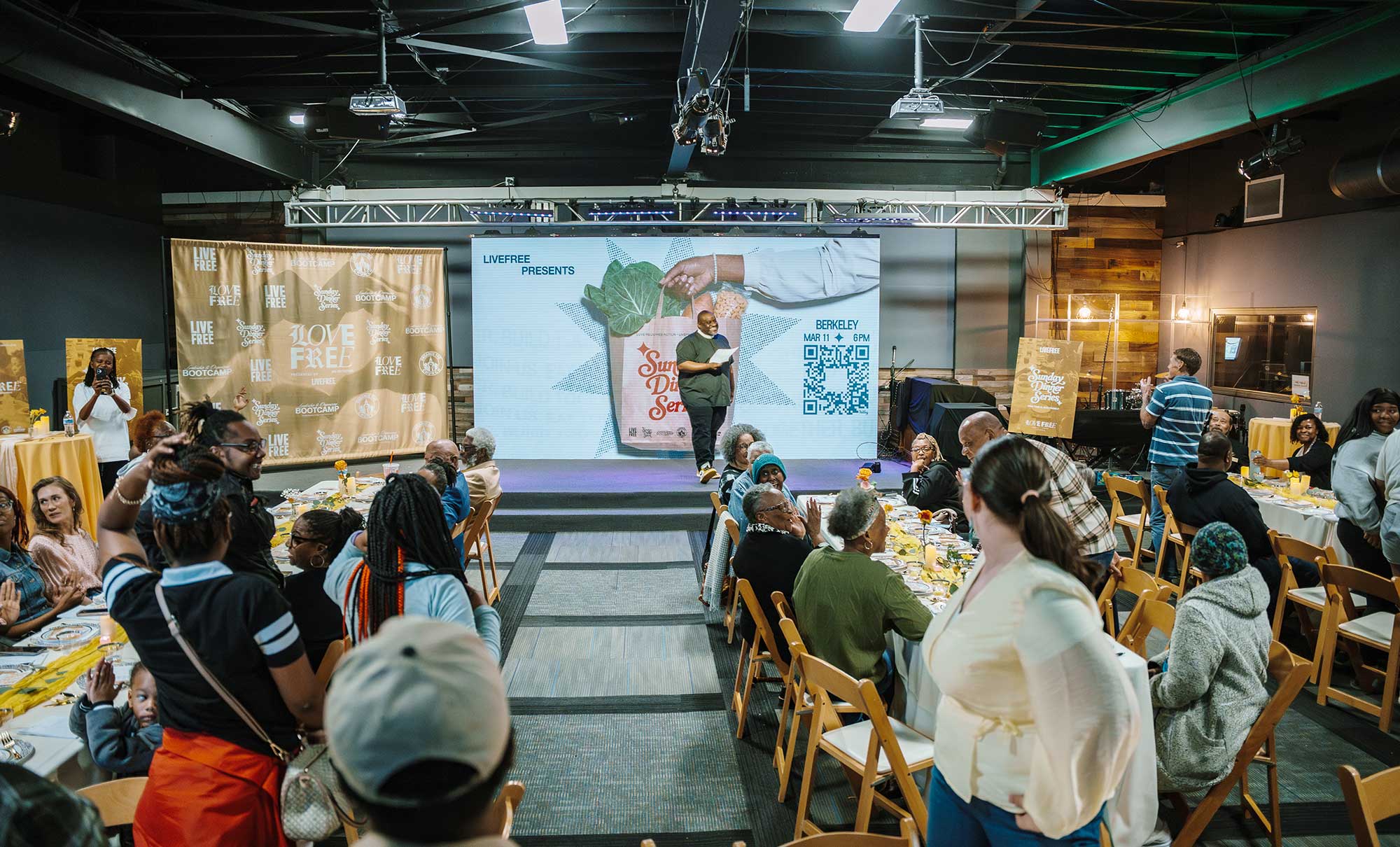 Pastor Mike, executive director of LIVE FREE USA, stands on stage in front of people sitting at tables as part of LIVE FREE’s Sunday Dinner series